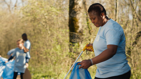 African american activist doing litter cleanup to fight illegal dumping, protecting the natural environment with garbage disposals. Woman working on collecting rubbish from the forest area. Camera A.の写真素材
