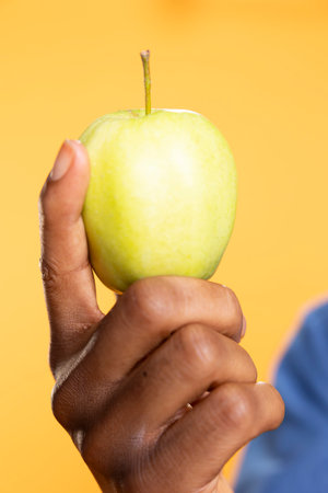 African american guy holding a green apple against yellow background, presenting local ethically sourced fresh produce in front of the camera. Zero waste enthusiast shows ripe fruits. Close up.の写真素材