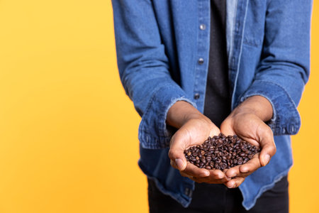 Coffee lover recommending black natural roasted beans against yellow background, promoting cold brew and caffeine drinks at a roastery. Freshness and aroma of delicious arabica seeds. Close up.の写真素材