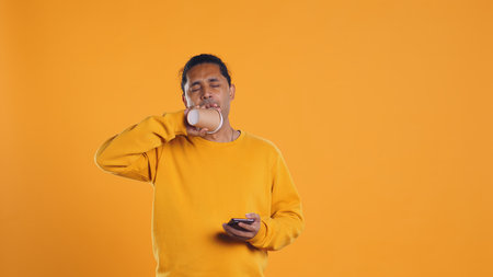 Indian man enjoying fresh coffee from disposable paper cup early in the morning to wake up and be energized. Person drinking hot beverage from recycled takeaway cup, studio background, camera Bの写真素材
