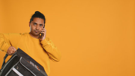 Man packing food in thermal backpack, fulfilling clients orders and answering phone ringing, studio backdrop. Indian person doing delivery to customers, discussing with them on cellphone, camera Aの写真素材