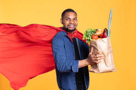 Superhero model with red cape posing with a bag of bio groceries, promoting sustainable lifestyle and locally grown veggies. Young person showcasing zero waste concept in studio.の写真素材