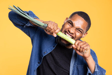 Portrait of young guy pretending to bite on a leek in the studio, posing against yellow background with organic fresh produce from farmers market. African american man promotes veganism.の写真素材