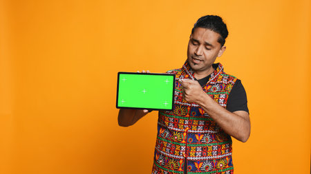 Portrait of indian man doing influencer marketing using green screen tablet, isolated over studio background. Smiling person holding empty copy space mockup device, camera Bの写真素材