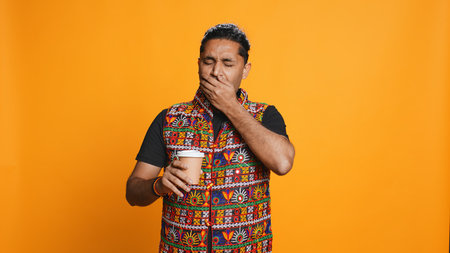 Tired indian man sipping fresh coffee from disposable paper cup early in the morning to wake up and be energized. Person drinking hot beverage from recycled takeaway cup, studio background, camera Bの写真素材