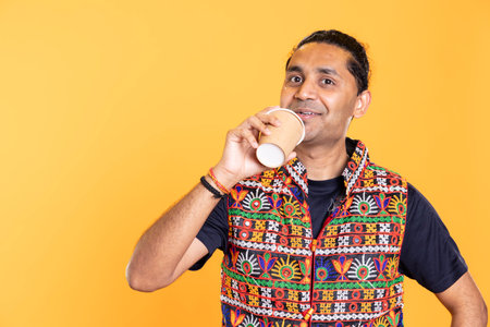 Portrait of cheerful man wearing traditional indian attire sipping fresh coffee to wake up and be energized. Smiling person with colorful vest drinking hot beverage, studio backgroundの写真素材