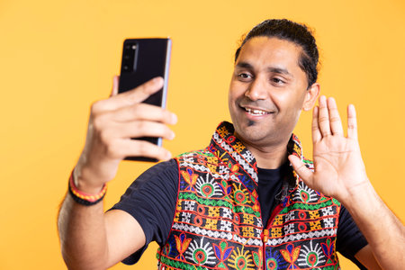 Joyous man doing waving hand gesture, talking with friend over internet videocall on smartphone. Upbeat indian person saluting mate during videoconference using phone, studio backgroundの写真素材