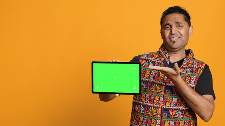 Upset indian man shows thumbs down sign gesturing holding isolated screen tablet, disapproving. Person does rejection hand gesture regarding mockup device, studio background, camera Aの写真素材