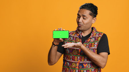Smiling indian man presenting cellphone with green screen display, isolated over studio background. Person in traditional clothing creating promotion with blank copy space mockup phone, camera Aの写真素材