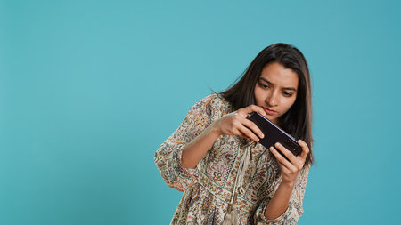 Happy indian woman entertained by videogames on smartphone, enjoying leisure time. Gamer enjoying game on phone, having fun defeating enemies, isolated over studio background, camera Aの写真素材