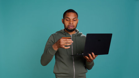 Happy man adding payment method on website using laptop, delighted to take advantage of promotions, studio background. Person thrilled to do internet shopping, typing credit card data, camera Bの写真素材