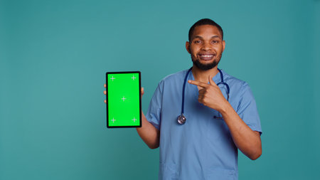 Portrait of male nurse showing medical instructions video on green screen tablet. Hospital employee holding chroma key device, isolated over blue studio background, camera Bの写真素材