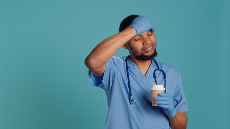 Tired healthcare specialist gaining energy with cup of coffee while at work, enjoying aroma. Fatigued medical practitioner drinking hot beverage, isolated over blue studio background, camera Aの写真素材