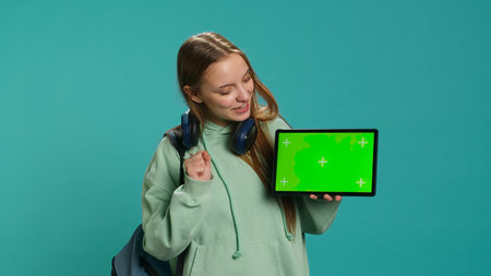 Portrait of joyous teenager reviewing tablet with mockup display, isolated over studio backdrop. Radiant young girl offering feedback on green screen digital device, camera Aの写真素材