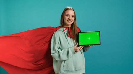 Joyous girl portraying superhero with red cape showcasing green screen tablet, studio backdrop. Joyful teenager posing as hero in costume on advertising chroma key digital device, camera Bの写真素材