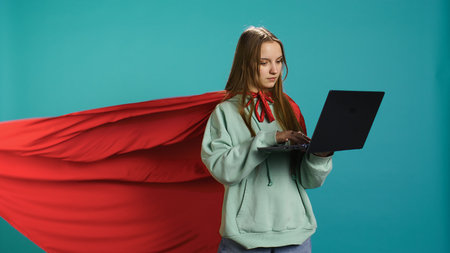 Young girl wearing superhero costume for Halloween working on laptop, isolated over studio background. Teenager dressed as comic book hero typing on notebook keyboard, camera Bの写真素材