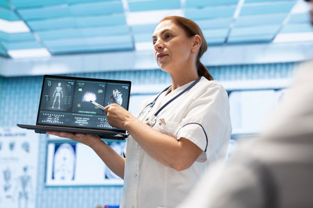 In a treatment center, medical experts team discuss patient care strategies. Licensed doctor pointing at heart and brain radiography scans on laptop, neurology and cardiology analysis.の写真素材