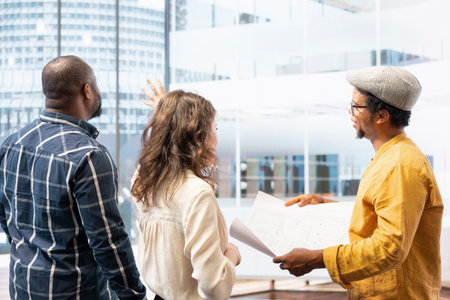 Clients and real estate agent on an office tour reviewing workspace design and blueprints, negotiating leasing contract for urban property. Planning corporate relocation in a building.の写真素材