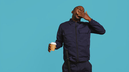 Tired security guard drinking coffee cup to stay awake, being tasked with preventing criminals. Portrait of weary bodyguard sipping caffeinated beverage during night shift, studio background, camera Bの写真素材