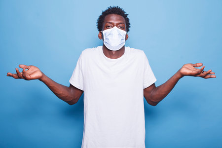 Portrait of African American man, looking bewildered, raises his shoulders while wearing face mask. Black guy stands against a blue background, gazing at camera with an uncertain expression.の写真素材