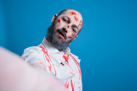 POV of actor dressed as thriller slasher movie zombie taking selfies between scenes on set. Man costumed as walking dead creature covered in blood makeup doing photos, isolated over studio backgroundの写真素材