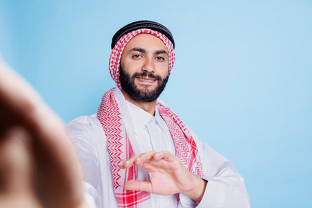 Smiling arab man wearing traditional attire holding mobile phone in arm and looking at front camera fpv. Muslim person with cheerful expression talking in videocall pov perspectiveの写真素材