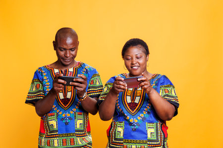 Smiling black man and woman dressed in ethnic clothes having online game competition on smartphone. Cheerful wife and husband couple playing videogame on mobile phone and having funの写真素材
