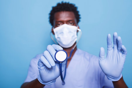 Portrait of black nurse using stethoscope to listen to heartbeats during cardiology checkup. Man with medical instrument, wears face mask and gloves to protect from coronavirus epidemic.の写真素材