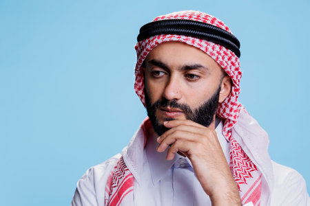 Thoughtful man wearing traditional muslim clothes rubbing chin and looking away. Arab person dressed in checkered ghutra headscarf standing in contemplation in studio on blue backgroundの写真素材