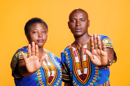 Serious black couple wearing ethnic clothes showing stop gesture and looking at camera. Confident...