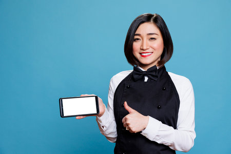 Cheerful asian woman receptionist holding smartphone with blank screen and showing thumb up portrait. Waitress standing with approval gesture and displaying mobile phone white touchscreenの写真素材