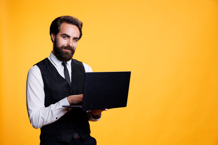 Young butler searches on internet for list of prices to put on menu, working to develop restaurant orders with pc against yellow background. Modern person holding device to check websites.の写真素材