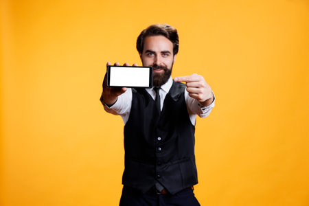 Skilled butler presents white blank display against yellow background, showing isolated mockup empty screen. Male waiter using smartphone with copyspace template in studio.の写真素材