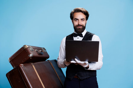 Doorkeeper holding modern laptop reflecting professionalism and competence, verifying list of bookings to maintain security and wellbeing. Young bellhop browses online platform.の写真素材
