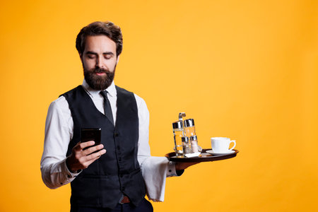 Professional butler checks smartphone and preparing to serve clients at table, carrying restaurant tray with cup of coffee. Luxurious stylish employee working in catering industry, uses moobile app.の写真素材