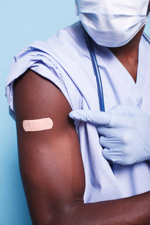 Close-up of African American nurse pointing to covid 19 immunization plaster while wearing gloves. Black individual with face mask motions to a bandage after receiving vaccination.の写真素材