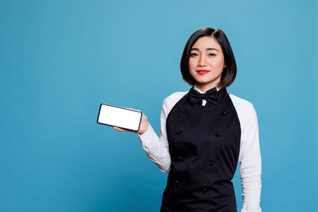 Asian waitress wearing restaurant uniform presenting smartphone with white screen for menu ads portrait. Young attractive woman receptionist showing mobile phone empty touchscreenの写真素材