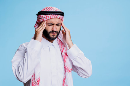 Muslim man wearing ghutra headscarf rubbing temples while having headache and high blood pressure. Arab person feeling pain and holding head while suffering from migraineの写真素材