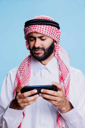 Arab man in islamic headscarf and thobe playing videogame on smartphone with tensed expression. Muslim gamer enjoying online gaming experience on mobile phone while posing in studioの写真素材