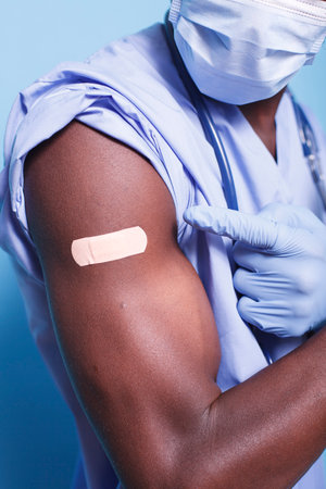 Close-up of African American gloved healthcare worker pointing to vaccine plaster to prevent and protect against COVID-19. Black person wearing face mask gestures to bandage on arm after vaccine shot.の写真素材