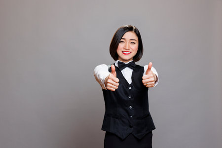 Smiling asian waitress with thumbs up recommending catering service portrait. Cheerful young attractive receptionist wearing restaurant uniform showing approval gesture and looking at cameraの写真素材