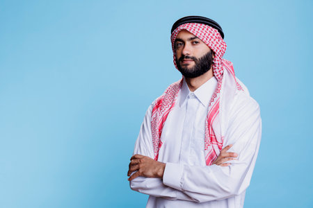 Confident muslim man dressed in traditional clothes standing with arms crossed portrait. Young arab man dressed in thobe and ghutra posing with folded hands and looking at cameraの写真素材