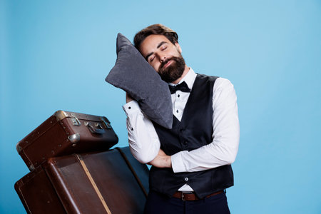 Tired bellboy posing with pillow on camera, expressing burnout and feeling sleepy at professional classy job. Hotel concierge taking quick nap after being overworked, exhausted doorman.の写真素材