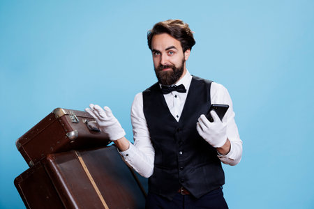 Professional bellman holds smartphone in hand and popsing next to trolley bags, portraying luxurious hospitality industry occupation. Young man in formal attire using mobile phone webpages.の写真素材