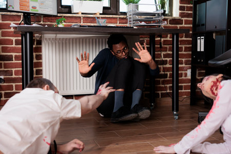 Actors playing zombies in horror movie scene grabbing BIPOC worker hiding under desk in office set. Man and woman with reanimated corpses roles in thriller film attacking victimの写真素材