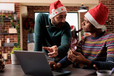 Diverse men coworkers wearing santa hats having conversation and brainstorming on laptop task in festive office. Colleagues discussing project in workplace during winter holidayの写真素材