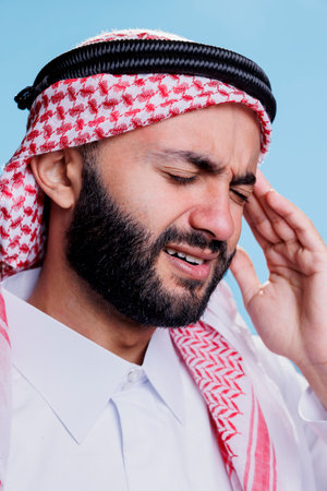 Arab man in pain wearing traditional clothes suffering from migraine. Muslim person dressed in islamic thobe and checkered ghutra headscarf rubbing temple while having headacheの写真素材
