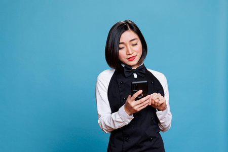 Smiling asian waitress wearing uniform texting on smartphone, managing catering service online. Restaurant woman receptionist typing message on mobile phone, posing in studioの写真素材