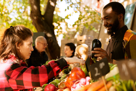 At a local farmers market, an African American man stands behind a stall selling fresh organic seasonal vegetables to a female customer. Young woman purchasing organic homegrown produce.の写真素材