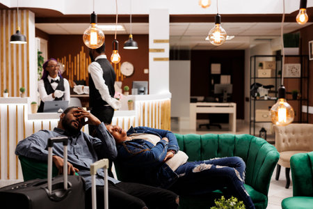 Jet lag prevention. Tired African American couple of tourists fall asleep after arrival to hotel, sleeping with luggage in lobby at night while waiting for check-in, having rest after long travelの写真素材
